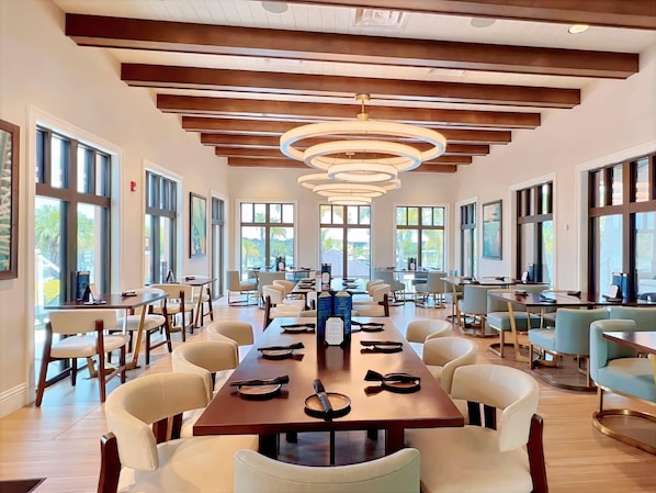 A spacious dining area with long tables, chairs, and large windows offering views of palm trees.
