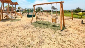 Un parque infantil con columpios y toboganes, rodeado de arena y follaje.