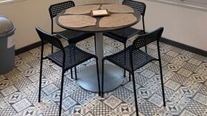 A dining area with a round table, four chairs, a trash bin, and a window with a view of greenery.