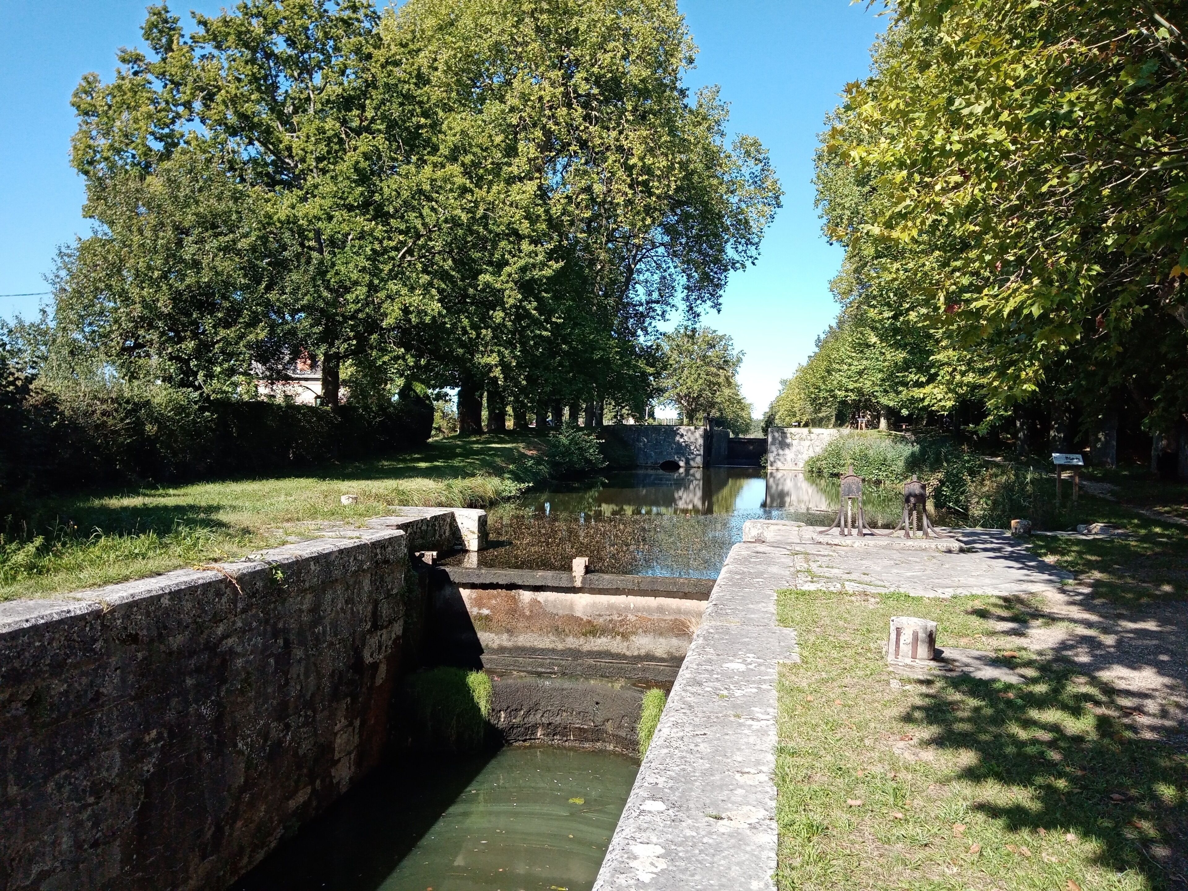 Een kanaal met sluisinstallatie, omgeven door bomen en grasvelden.