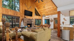 A cozy cabin interior with a stone fireplace, wooden furniture, and large windows offering a view of the forest.