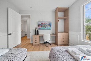 Quarto moderno com cama, escrivaninha e cadeira. Há um quadro na parede e uma janela com vista para árvores.