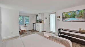 A modern, well-lit room with a bed, a desk, a chair, a small kitchenette, and a TV displaying a tropical pool view.