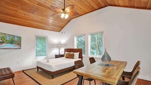Una camera da letto con soffitto in legno, un letto, un comodino, una lampada, un tavolo da pranzo con sedie e una finestra con persiane.