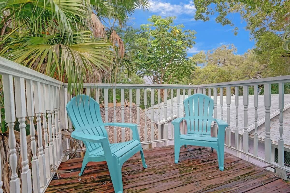 A wooden deck with two turquoise Adirondack chairs, surrounded by a white picket fence and lush greenery.