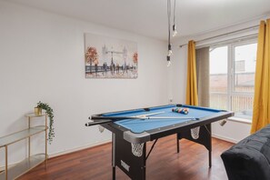 A pool table in a room with a window, a painting of a bridge, and a plant on a shelf.