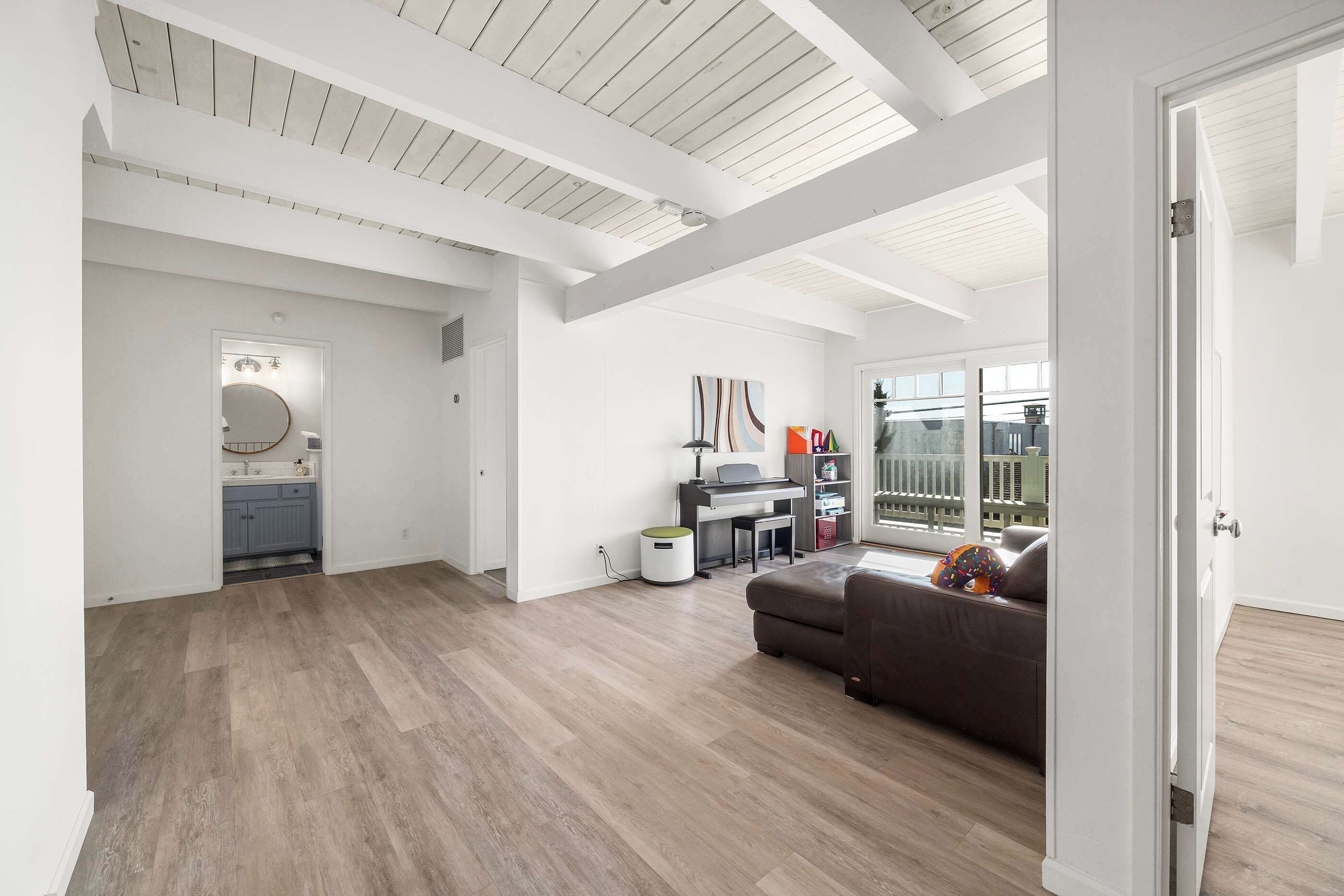 A spacious living area with a wooden floor, a sofa, a desk with a computer, and a view through a glass door.