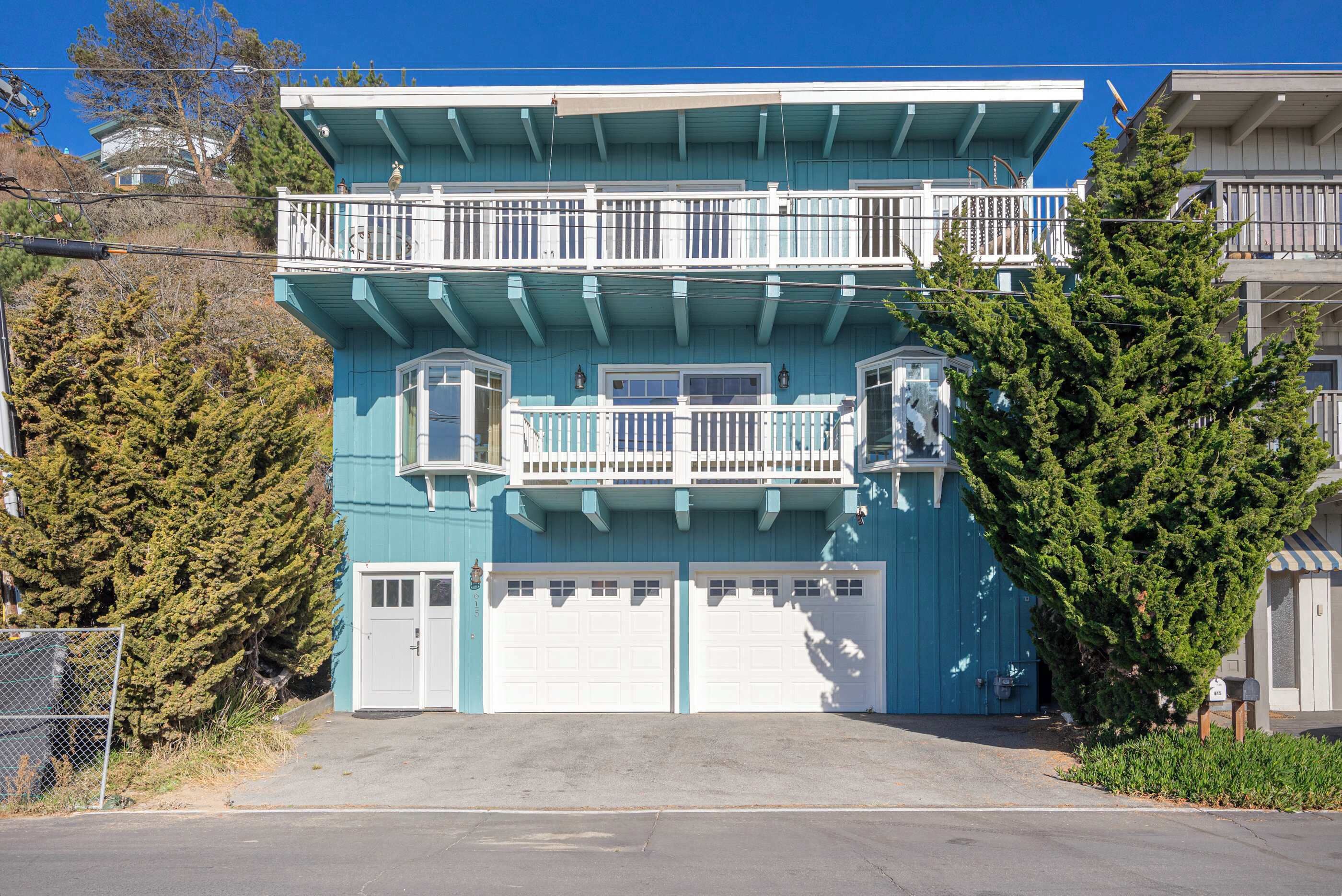 Un bâtiment à deux étages, avec une façade bleue, des portes de garage blanches et un balcon à l’étage supérieur.