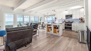 Een ruime woonkamer met een bank, eettafel en keuken met witte kastjes en een houten plafond.