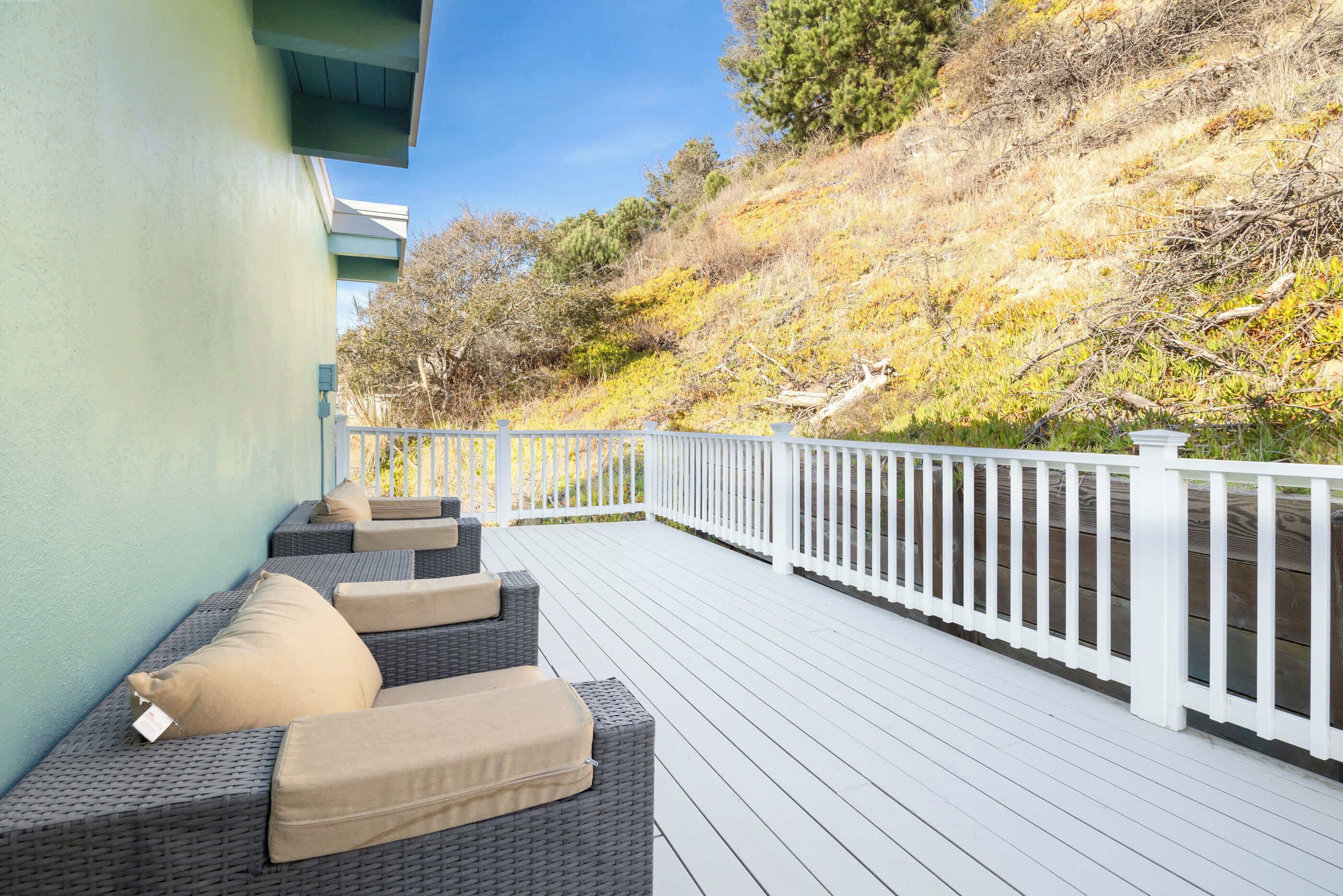 A covered outdoor seating area with wicker furniture and beige cushions, a white railing, and a hillside backdrop.