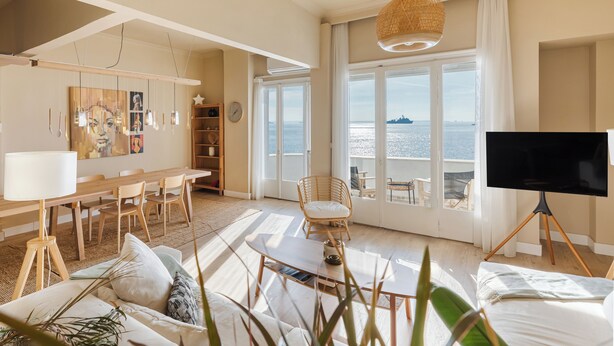 A spacious living room with a large window overlooking a body of water, a dining area with wooden furniture, and a television on a tripod stand. - Infinity & Beyond Seafront Apartments by Halu! (Thessaloniki)