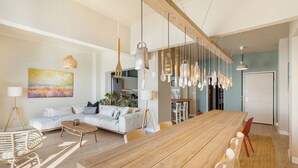 A spacious living room with a wooden dining table, a white sofa, and hanging pendant lights. - Infinity & Beyond Seafront Apartments by Halu! (Thessaloniki)