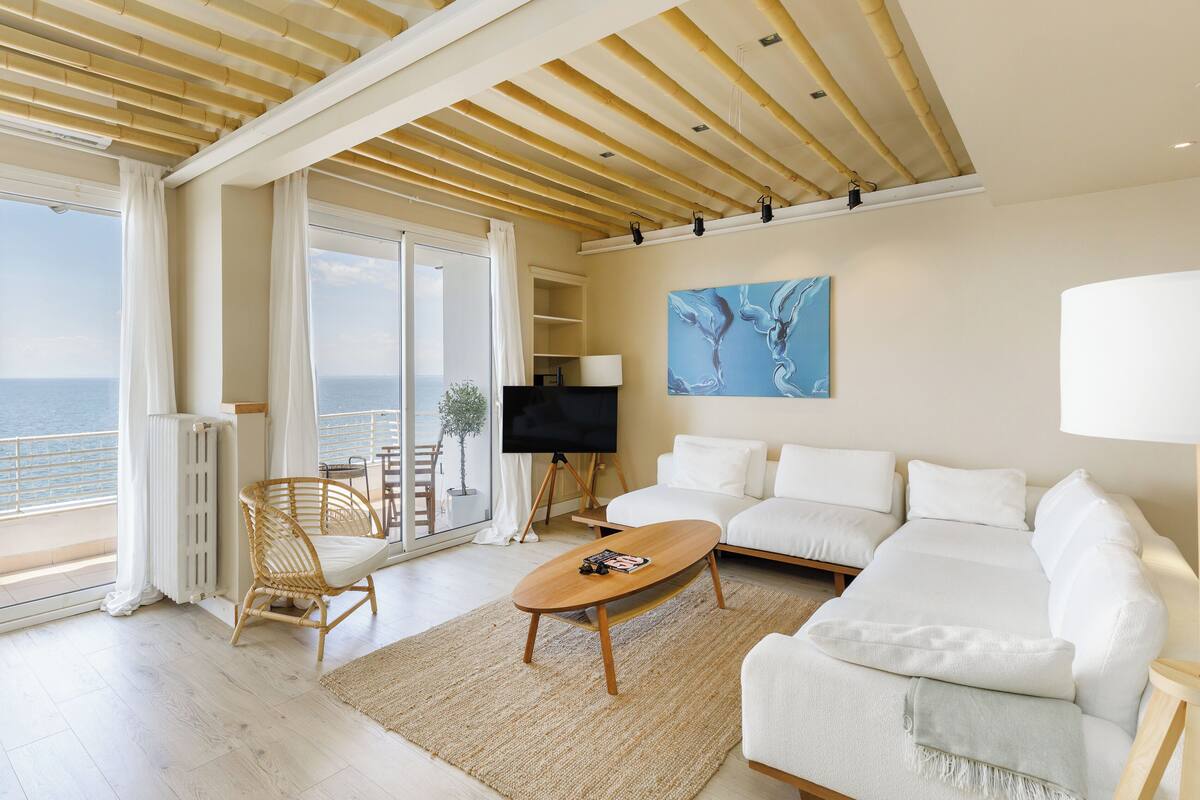 A spacious living room with a large sofa, a coffee table, and a TV. There is a balcony with a view of the sea.