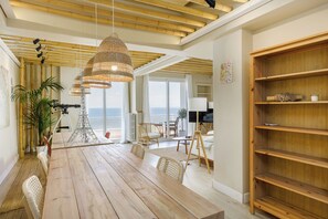 A spacious dining area with a wooden table and chairs, a large woven pendant light, and a view of the sea through large windows. - Infinity & Beyond Seafront Apartments by Halu! (Thessaloniki)