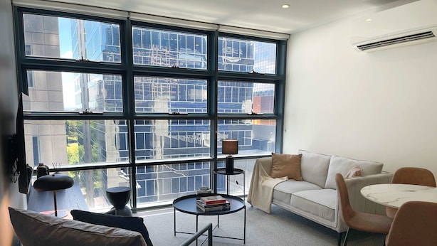 A modern living room with a large window, a sofa, a coffee table, and a dining area. - Modern and Elegant 1BD Apartment in the CBD (Melbourne)
