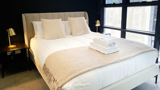 Un lit bien fait, avec une couette blanche, un plaid beige et des serviettes pliées posées sur le lit. On y trouve deux tables de chevet avec des lampes, ainsi qu’une grande fenêtre donnant sur des bâtiments et de la verdure.