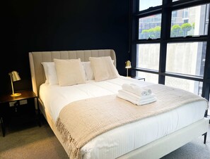 Una cama bien hecha con edredón blanco, una manta beige y toallas enrolladas sobre la cama. Hay dos mesitas de noche con lámparas y un ventanal con vistas a edificios y zonas verdes.