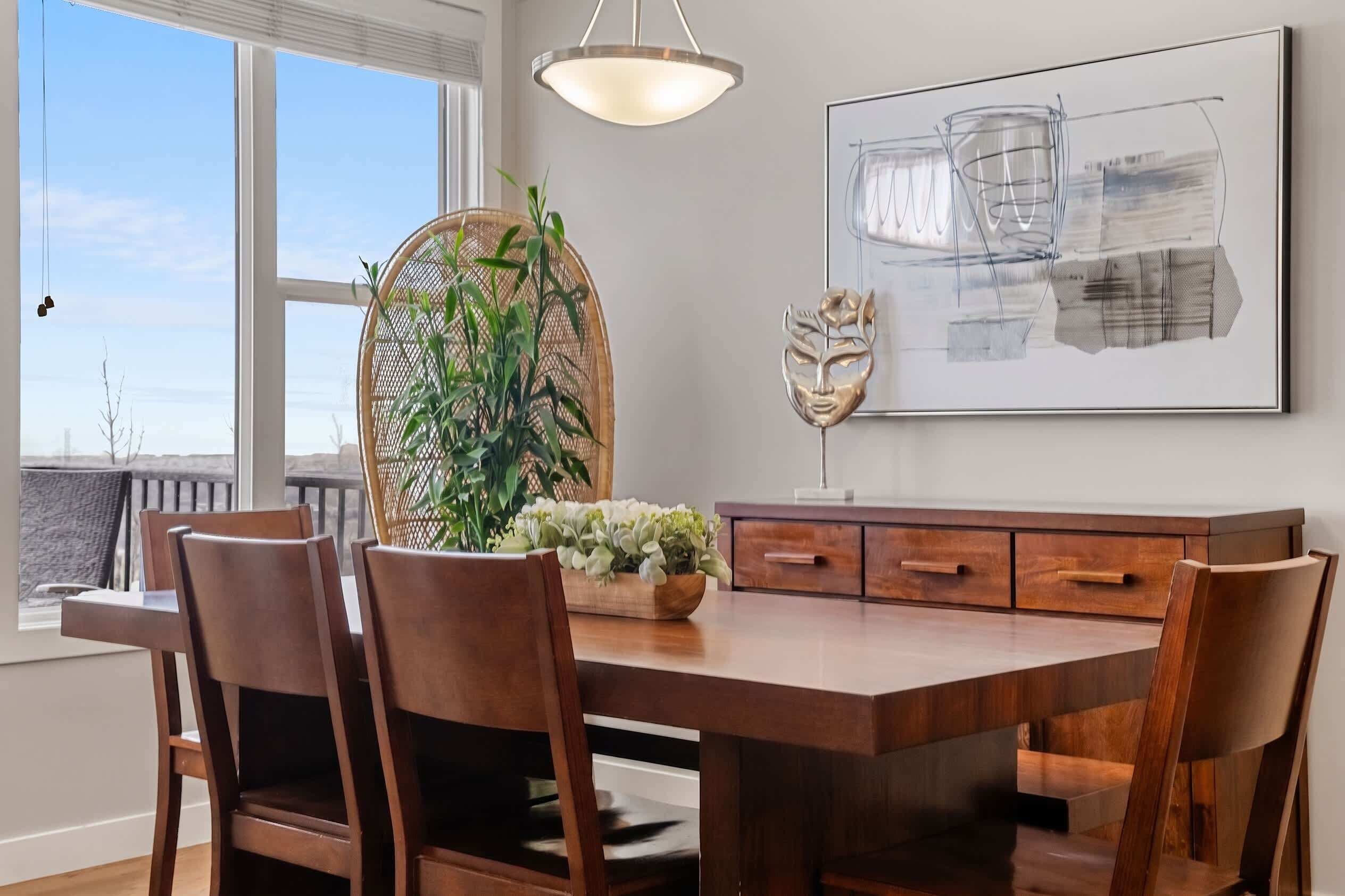 A dining area with a wooden table, chairs, a hanging light fixture, a wall-mounted abstract painting, and a potted plant.
