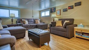 A living room with a sectional sofa, ottoman, and a coffee table. There are two windows providing natural light, and the room has a wooden floor. - LOOK!  Spacious Home in the Heart of Hilo - Bonus Rec Room! (Hilo)