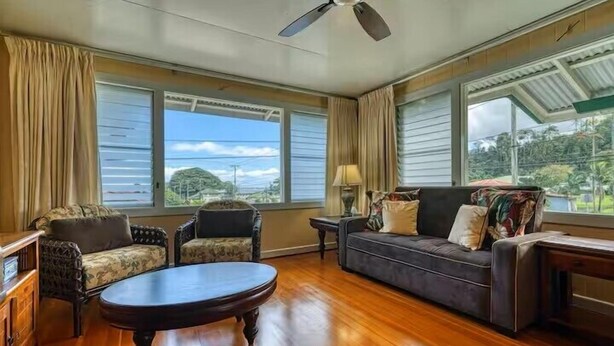 A spacious living room with a wooden floor, a ceiling fan, and large windows offering a view of the outdoors. - LOOK!  Spacious Home in the Heart of Hilo - Bonus Rec Room! (Hilo)