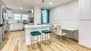 A modern kitchen with white cabinetry, a central island with green stools, a dining area with a white table and chairs, and a window with a blue curtain. - 6 Mi to Eagles Games: Stylish Philly Townhome! (Philadelphia)