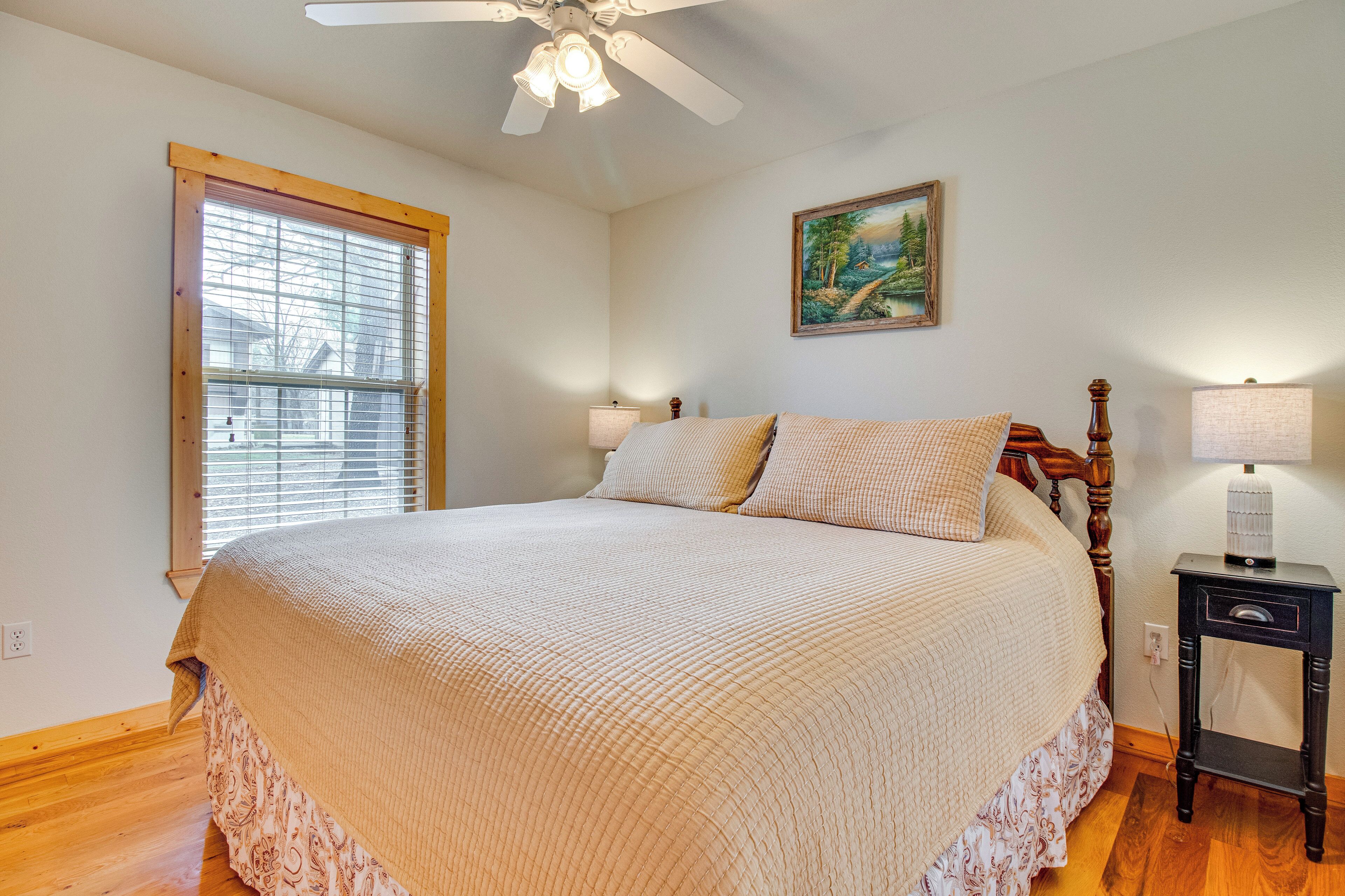 A bedroom with a large bed, a ceiling fan, a window with blinds, a nightstand with a lamp, and a painting on the wall.