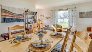 A dining area with a wooden table set for four, surrounded by wooden chairs. There is a bookshelf with books and decorative items, a sofa, and a window with curtains. - 17 Mi to Brookville Lake: Home w/ In-unit Laundry (Connersville)