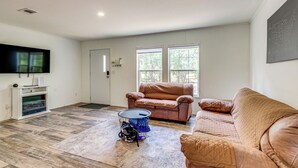 A spacious living room with hardwood flooring, a large brown leather couch, a smaller brown armchair, and a flat-screen TV mounted on the wall.
