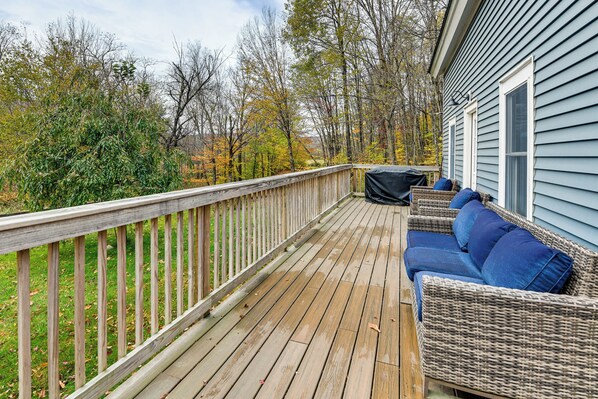 A wooden deck with a wicker sofa and blue cushions, surrounded by trees and a house. - Snow Days & Ski Getaways: Belvidere Farmhouse (Belvidere)