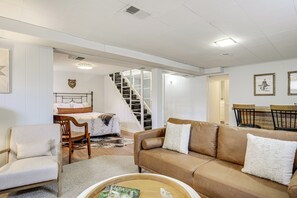 Una sala de estar con un sofá marrón, una silla de madera, una mesa de comedor con sillas, una escalera y una cama con cabecera.