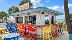 Un bar frente a la playa con sillas y mesas de colores, un cartel de menú y un letrero que dice 'The Bar'.