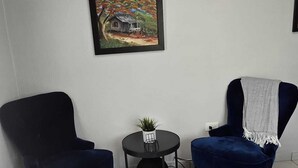Ein kleiner, sauberer Raum mit zwei dunkelblauen Sesseln, einem kleinen runden Tisch mit einer Pflanze, einem gerahmten Gemälde an der Wand und einem Bett mit einer gestreiften Decke.
