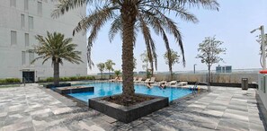 Una piscina en la azotea con sillones de descanso, palmeras y un edificio moderno al fondo.
