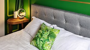 A bedroom with a green wall, a painting, a bedside table with a lamp, and a bed with white bedding and a green pillow.
