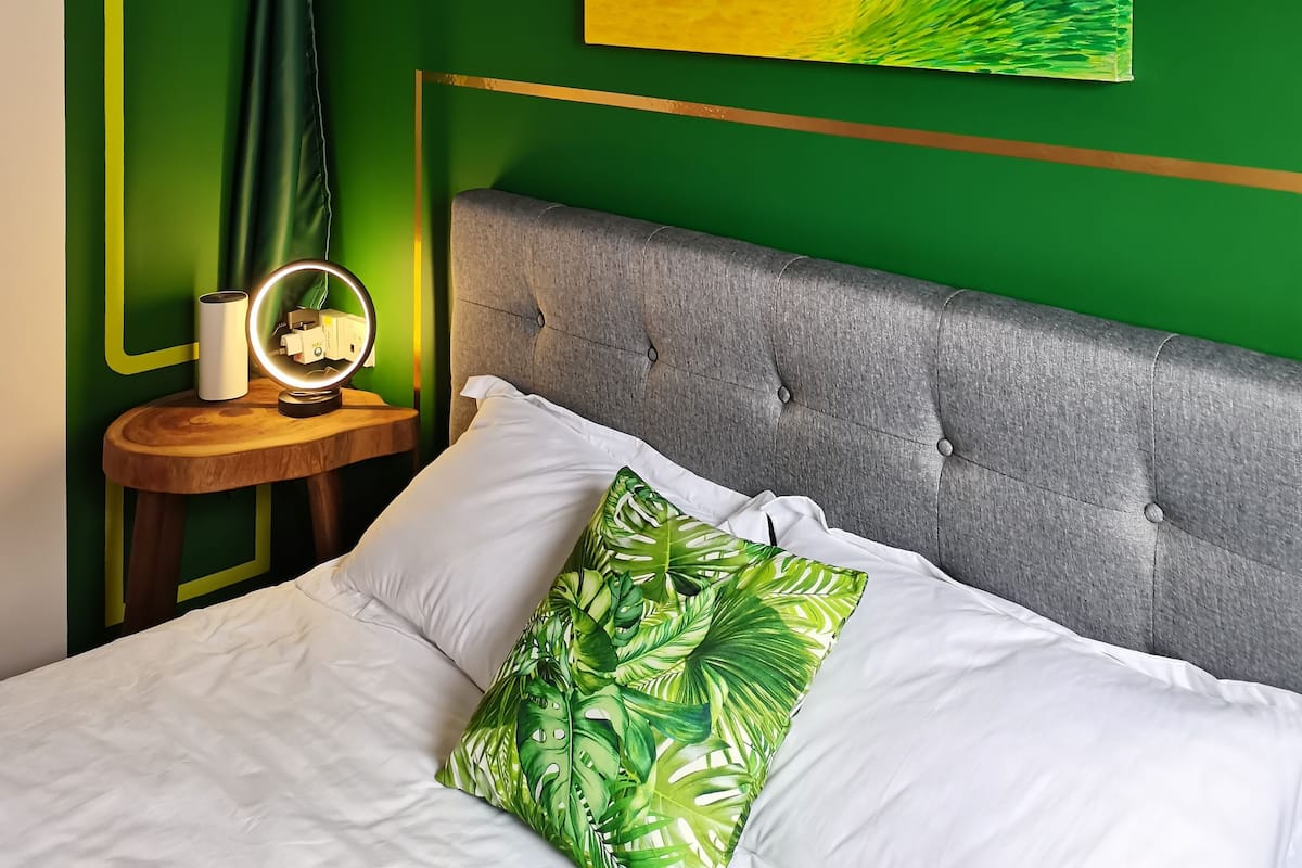 Ein Schlafzimmer mit grüner Wand, einem Gemälde, einem Nachttisch mit Lampe und einem Bett mit weißer Bettwäsche und einem grünen Kopfkissen.