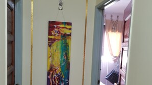 A hallway with a painting on the wall, a door, and a glimpse of a bedroom with a bed and curtains.