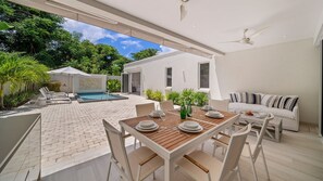 An outdoor dining area with a pool, white furniture, and a view of the surrounding greenery. - Porters Place 18 4 Bedroom Villa (Holetown)