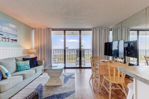 A modern living room with a sofa, coffee table, and a dining area with chairs and a TV. - 0207 Dancing Tides 1 Bedroom Condo (Carolina Beach)