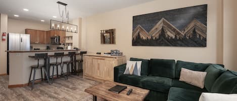 A modern living room with a green sofa, a coffee table, and a kitchen area with a bar and stools.