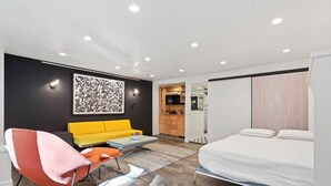Ein modernes Hotelzimmer mit einem Bett, einem gelben Sofa, einem roten Sessel, einem kleinen Tisch und einer Küchenzeile im Hintergrund.