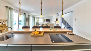 Eine moderne Küche mit Kochinsel, integriertem Backofen und Spüle. Es gibt Pendelleuchten, einen Essbereich mit Sofa und Stühlen sowie eine Treppe zu einer weiteren Ebene.