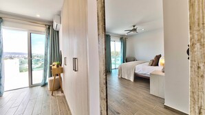 Ein modernes Hotelzimmer mit einer großen Schiebetür, die auf einen Balkon führt, einem Bett mit weißer Bettdecke, einem Nachttisch mit Lampe und Blick auf die Stadtlandschaft.