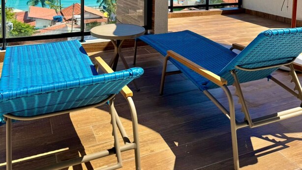 Un balcón con dos sillones azules, una pequeña mesa redonda y una sombrilla beige. El balcón da a un océano azul cristalino con palmeras y edificios al fondo.