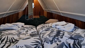 Una habitación tipo tienda de campaña, estrecha y triangular, con dos camas, una mesita y una lámpara en el techo.