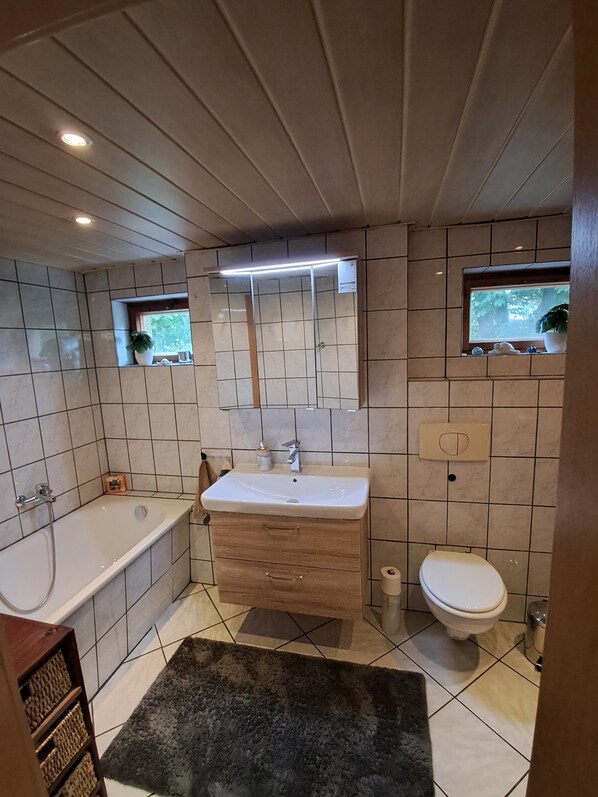 A bathroom with a bathtub, sink, and toilet. There are two windows with plants, a mirror above the sink, and a wooden ceiling.