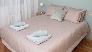 Una cama bien hecha con dos toallas dobladas, una lámpara de noche y una ventana con cortinas translúcidas.