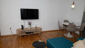 Una sala moderna con televisor de pantalla plana, una mesa redonda, un sofá azul verdoso y una zona de comedor con una mesa blanca y sillas grises.