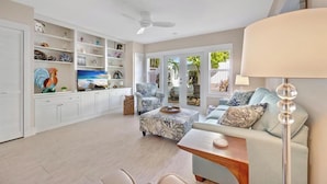 Un salón amplio con una televisión grande, un ventilador de techo y vistas al exterior a través de amplias ventanas.