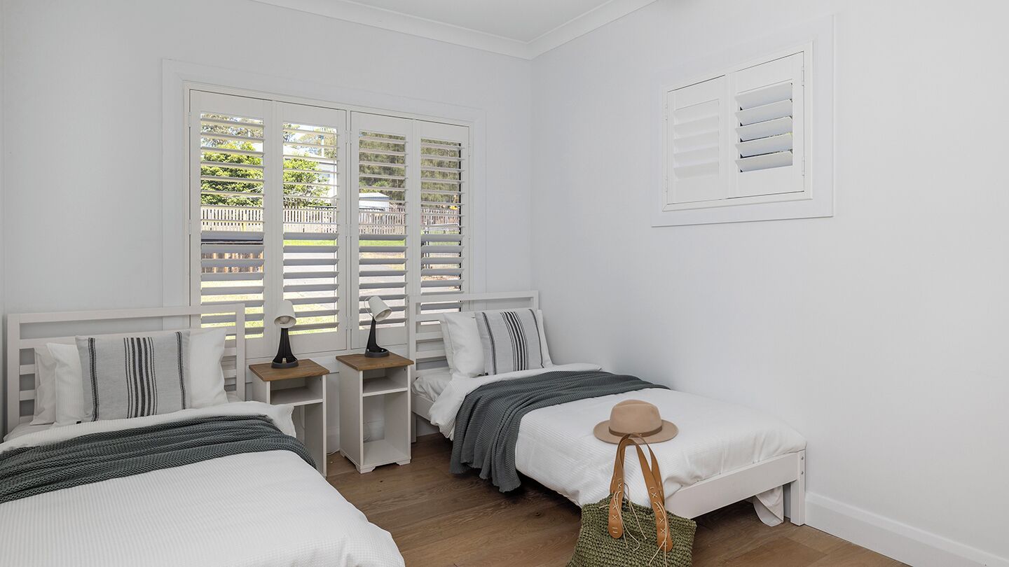 A bedroom with two beds, a window with shutters, a bedside table with a lamp, and a wall-mounted air vent.