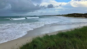Une plage avec des vagues, des dunes herbeuses et un ciel nuageux.